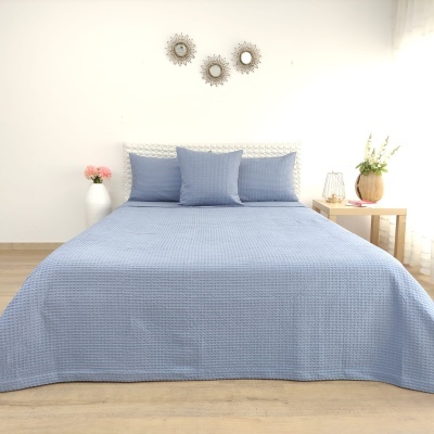 Cama de casal com colcha azul texturizada e almofadas, mesa de cabeceira com vaso, jarra de flores e três espelhos redondos na parede.