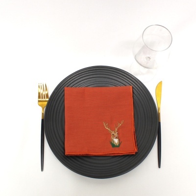 Conjunto de jantar com prato preto, guardanapo castanho com bordado de veado, talheres dourados e copo transparente.