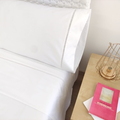 Lençóis brancos com costura decorativa e mesa de cabeceira com livro e luminária