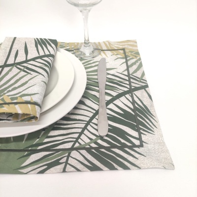 Individual de mesa com padrão de folhas verdes e guardanapo combinado, pratos brancos, faca de metal e taça de vidro.