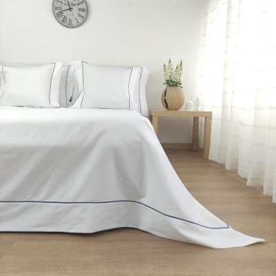 Cama com roupa branca com linha azul, relógio de parede, mesa de madeira com vaso de flores.