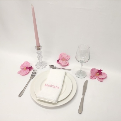 Conjunto de mesa com pratos brancos, talheres em metal, copo de vidro, guardanapo bordado, vela rosa e flores cor-de-rosa