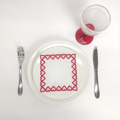 Mesa posta com prato branco, guardanapo com corações vermelhos, talheres metálicos e copo com base vermelha.