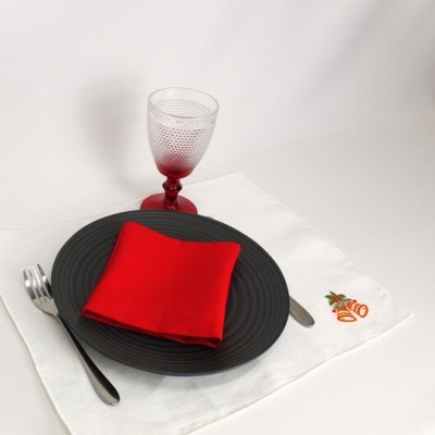 Mesa posta com prato preto, guardanapo vermelho, talheres metálicos, copo de vidro com pé vermelho e toalha branca bordada