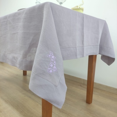 Toalha de mesa cinzenta com bordado de coelho lilás sobre mesa de madeira