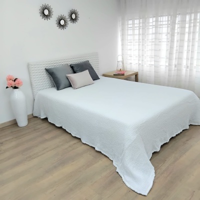 Quarto com cama de casal com colcha branca, almofadas cinza e rosa, mesa de cabeceira e vaso com flores.