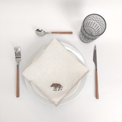 Mesa posta com talheres de cabo de madeira, guardanapo bege com ornamento de elefante, prato branco transparente e copo de vidro decorado.