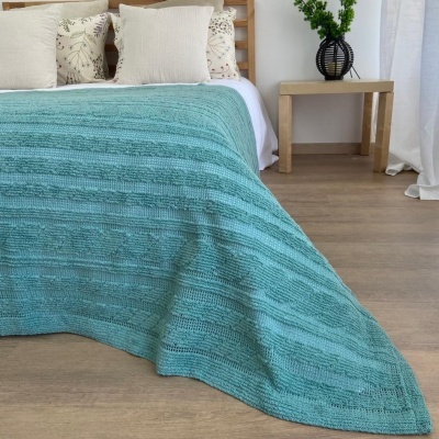 Colcha azul de malha numa cama com almofadas e mesa lateral com vaso preto e planta