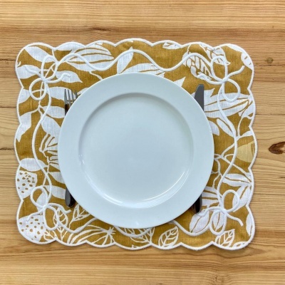 Prato branco redondo sobre guardanapo amarelo com padrão de folhas brancas e bordas recortadas numa mesa de madeira clara.