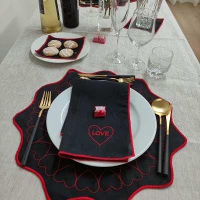 Mesa posta com toalha bege, placemats e guardanapos pretos com bordas vermelhas, pratos brancos, talheres dourados e copos de vidro