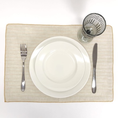 Mesa posta com jogo americano bege, pratos brancos, talheres prateados e copo transparente com padrão preto