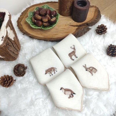 Quatro guardanapos brancos bordados com animais sobre superfície branca com castanhas, pinhas, velas e tábua de madeira