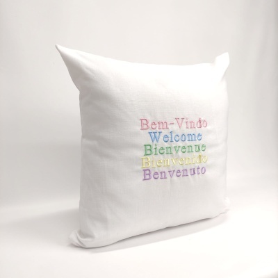 Almofada branca com bordado colorido de palavras de boas-vindas em várias línguas