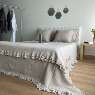 Cama com colcha bege clara com babados, parede branca com decorações hexagonais pretas, piso em madeira clara e mesa com vaso.