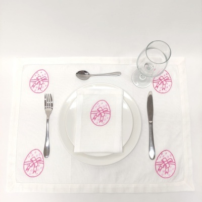 Jogo americano branco com bordado rosa de ovos de Páscoa, prato branco, guardanapo branco, talheres metálicos e taça de vidro
