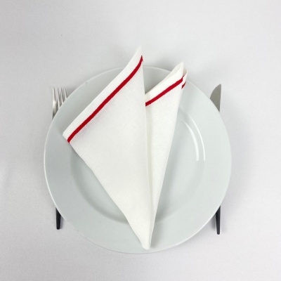 Conjunto de jantar com prato branco, guardanapo branco com risca vermelha, garfo e faca numa toalha branca