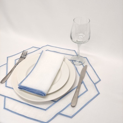 Pratos brancos, guardanapo com borda azul, talheres metálicos e copo de vinho em fundo branco.