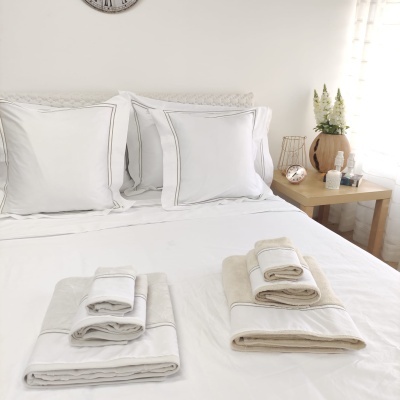 Conjunto de cama branco com almofadas e lençóis e conjuntos de toalhas brancas e bege sobre cama ao lado de mesa com vaso e flores.