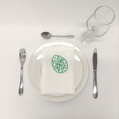 Mesa posta com prato branco, guardanapo com bordado verde, talheres de metal e copo de vidro