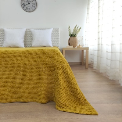 Cobertor amarelo texturizado numa cama branca com almofadas, relógio e mesa de apoio