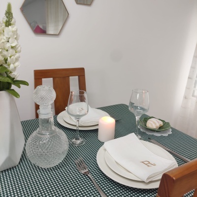 Mesa de jantar com toalha xadrez, pratos brancos, guardanapos com 'R', garrafa de vidro, copos de vinho, vela e flores brancas