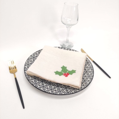 Conjunto de prato cerâmico azul e branco com guardanapo bege decorado com azevinho, talheres pretos e dourados, e copo de vidro transparente