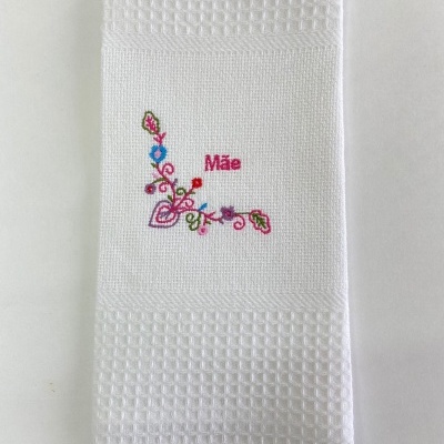 Pano de cozinha branco com bordado colorido e a palavra 'Mãe'.