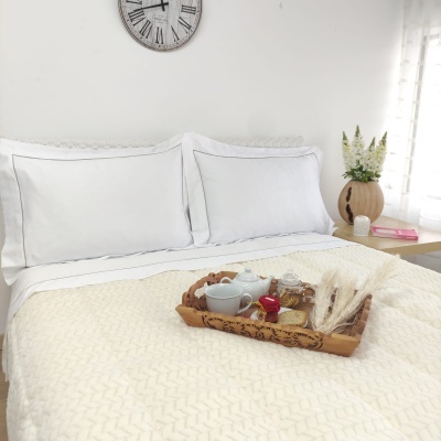 Cama de casal arrumada com roupa branca e edredão creme texturizado, bandeja de pequeno almoço em madeira, relógio de parede e vaso com flores ao lado.