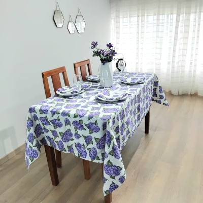 Mesa de madeira com toalha floral lilás e verde, arrumada com pratos e decoração floral.