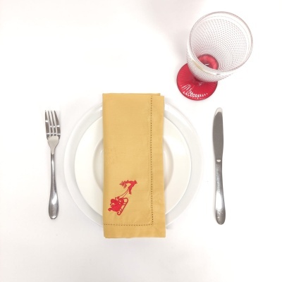 Mesa posta com prato branco, talheres metálicos, guardanapo amarelo bordado vermelho e copo transparente em base vermelha.