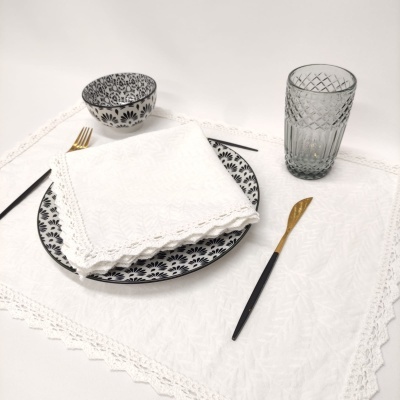 Conjunto de mesa com prato e tigela de porcelana, guardanapo branco bordado, talheres pretos e dourados, copo cinza com relevo e toalha branca bordada.
