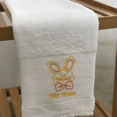 Toalha branca com bordado de coelho e texto 'Feliz Páscoa' pendurada em estrutura de madeira.