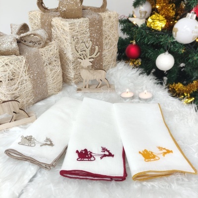 Três panos de cozinha brancos com bordados de trenó e rena em cores diferentes, junto de velas, presentes rústicos e árvore de Natal.
