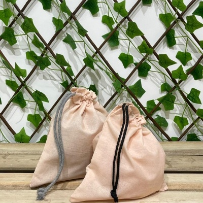 Dois sacos de pano bege com cordões, sobre superfície de madeira, frente a treliça com folhas verdes