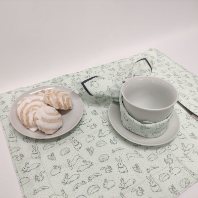 Chávena e pires brancos em cima de guardanapo verde claro com desenhos de animais, faca ao lado e prato com bolachas açucaradas