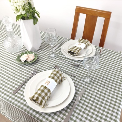 Mesa posta com toalha xadrez verde, prato branco, guardanapo xadrez com anel branco e R, copos de vidro, jarra branca com flores e macarons