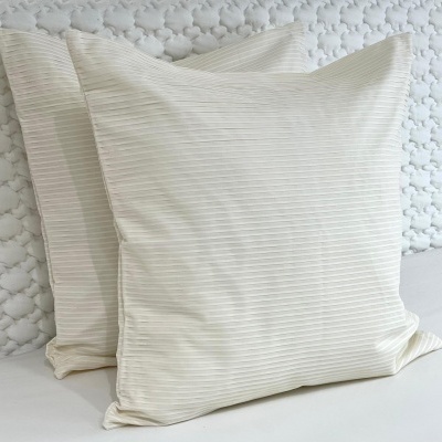 Duas almofadas quadradas creme com padrão de linhas finas paralelas, contra parede texturizada cinza claro.