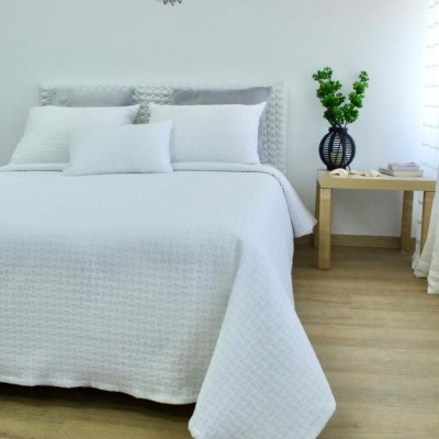 Cama de casal com colcha branca e almofadas, mesa de cabeceira com vaso de plantas e cortinas brancas