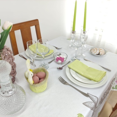 Mesa posta com pratos brancos, guardanapos verdes, talheres, copos, castiçais com velas verdes, cesta amarela com ovos e doces em taça.