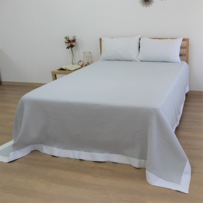 Cama com colcha cinza e almofadas brancas numa divisão com chão de madeira e móvel lateral