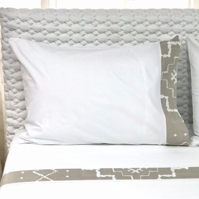 Travesseiro branco com borda cinza e padrão geométrico branco numa cama com roupa de cama combinando e cabeceira cinza