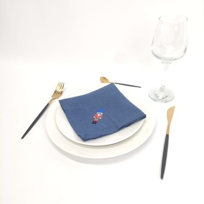 Conjunto de mesa com prato branco, guardanapo azul com bordado, talheres preto e dourado, copo de vidro