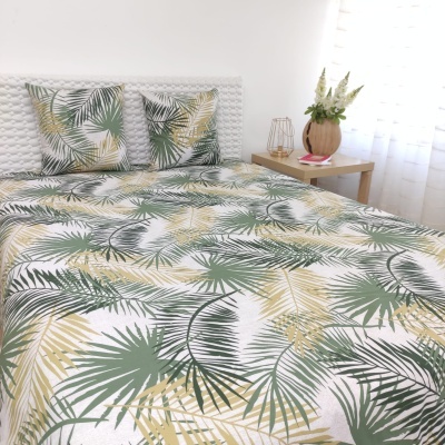 Cama com roupa de cama branca com padrão de folhas tropicais em verde e amarelo, almofadas combinando, mesa lateral de madeira com vaso e flores, ambiente iluminado por cortinas brancas.