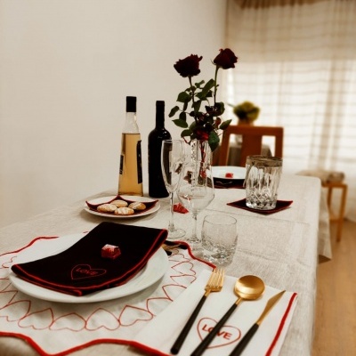 Mesa posta com pratos brancos, guardanapos pretos bordados em vermelho, talheres dourados e pretos, copos e taças, garrafas de vinho, rosas vermelhas em vaso, cortinas ao fundo