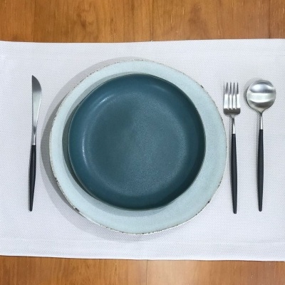 Conjunto de loiça com pratos azul esverdeado e cinza, talheres com cabo preto, sobre placemat branco e mesa de madeira clara
