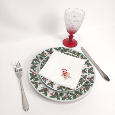 Conjunto de pratos com padrão de azevinho, guardanapo branco com boneco de gengibre, talheres prateados e copo vermelho