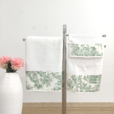 Conjunto de toalhas brancas com padrão verde penduradas num suporte metálico ao lado de vaso branco com flores cor-de-rosa.