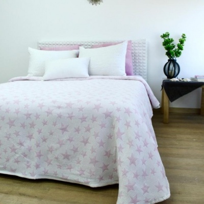 Cama de casal com colcha rosa clara estampada e cabeceira branca rattan, mesa de cabeceira com vaso e planta verde, chão de madeira e cortina branca