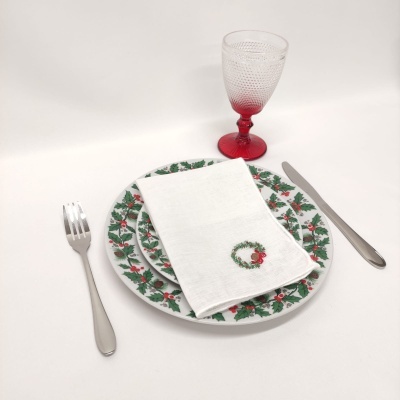 Mesa posta com pratos decorados, guardanapo branco bordado, talheres metálicos e copo vermelho texturizado