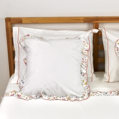 Almofada branca com folhos floridos e bordas vermelhas numa cama com cabeceira de madeira